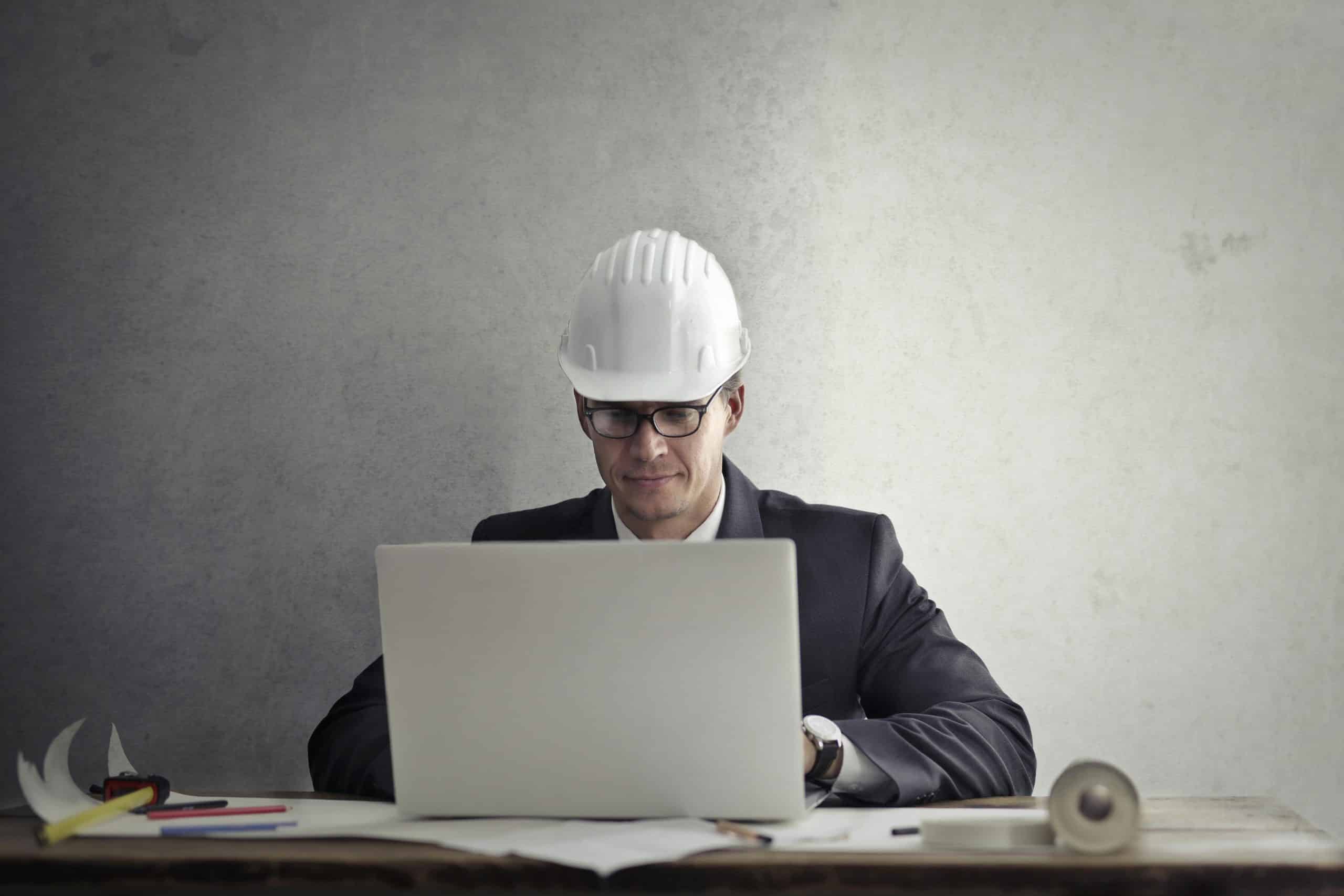 Professional engineer wearing hardhat working on construction plans at desk with laptop in office.